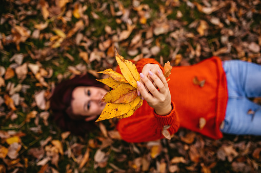 Beautiful young woman lying on autumn leaves in park and smiling. Holding some leaves in the hands.