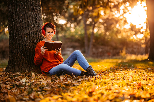 Beautiful young woman leaning on a tree in the park in autumn, listening to music via headphones and reading a book.