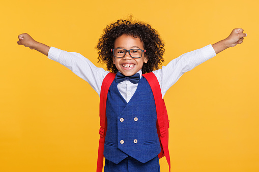 Excited african american boy in school uniform and glasses  raising arm and smiling while celebrating success against yellow backdrop