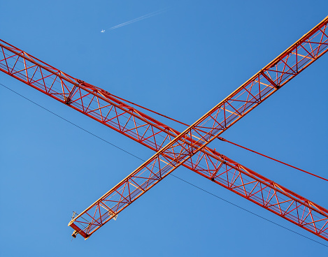 a perspective view of two orange crane booms crossing each other in front of deep blue sky with a passing airplane