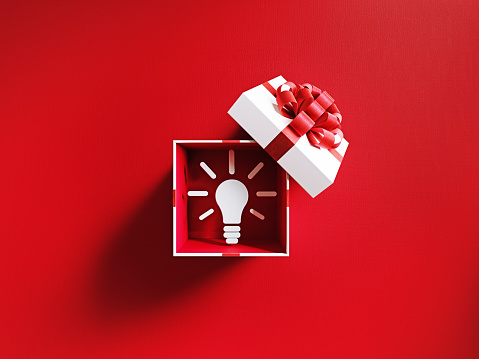 Lightbulb symbol is coming out of a white gift box tied with red ribbon on red background. Horizontal composition with copy space. Directly above. Great use for gift idea concepts.