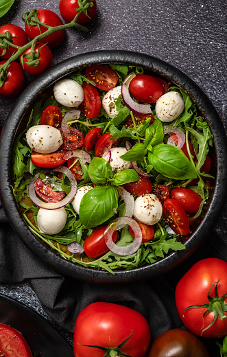 Salad with traditional italian mozzarella cheese with arugula and tomatoes on dark concrete table with cooking ingredients cherry tomatoes, basil. Top view.