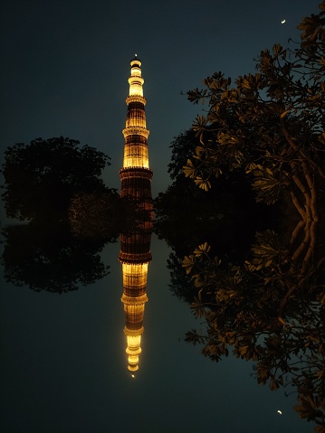 A vertical shot of the Qutb Minar complex and its reflection in the water at night in Delhi, India