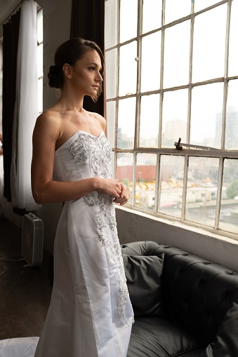 A vertical shot of a Caucasian woman in a beautiful wedding gown looking out of the window