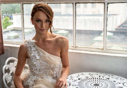 A beautiful Caucasian woman in a white one-shoulder wedding dress sitting on a chair