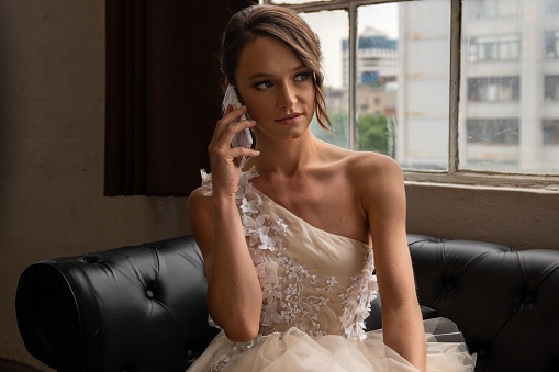 A Caucasian woman in a white wedding dress sitting in a couch and talking on the phone