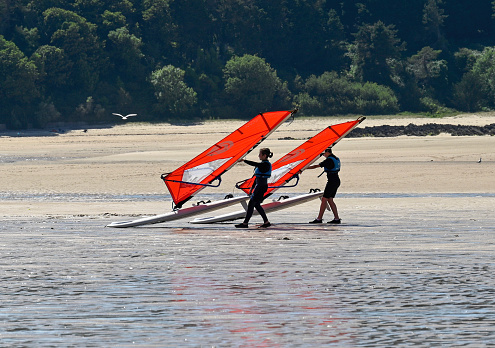 Erquy, France, July 5, 2022 - Two young people pull their windsurfing board into the water on the beach of Erquy