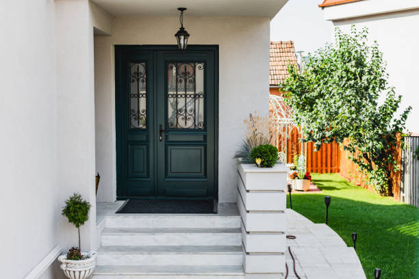 Front side of a home with a green wooden door and white stairs next to terrace with grass. A lamp is hanging in front of the door and patio is decorated with a small tree in a pot