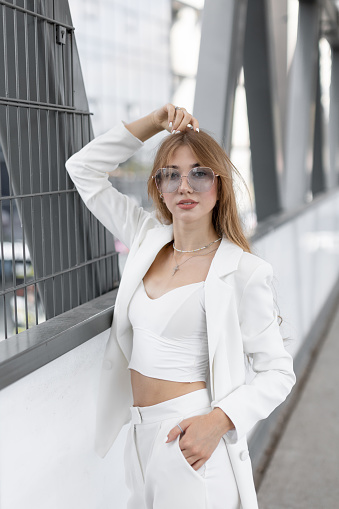 Stylish adorable pretty woman with wavy hair wearing white stylish suit and sunglasses standing and having fun in the street.