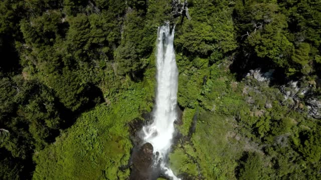 Aerial dolly in lowering on Salto El Leon waterfall falling into rocky pool surrounded by green woodland, Pucon, Chile