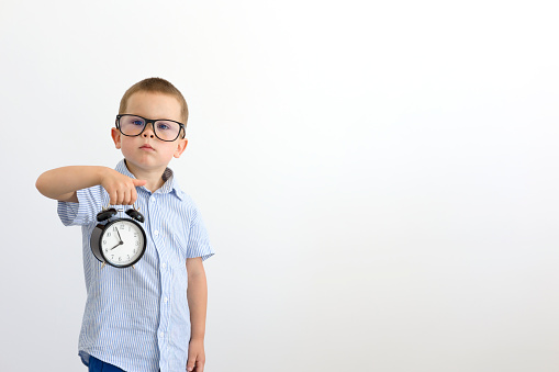 A boy in a blue shirt holds an alarm clock and looks straight ahead. Sad little boy. Back to school. Child on a light background with space for copy. School, student, learning. High quality photo