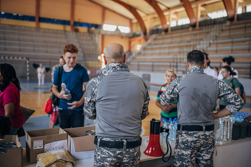 Diverse group of people, soldiers on humanitarian aid to civilians in school gymnasium, after natural disaster happened in city.