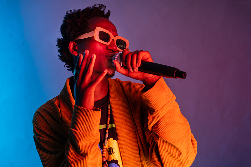A happy smiling African-American man singing into a microphone and gesturing with his hand, he is wearing a smart jacket and sunglasses with a gradient blue to purple background