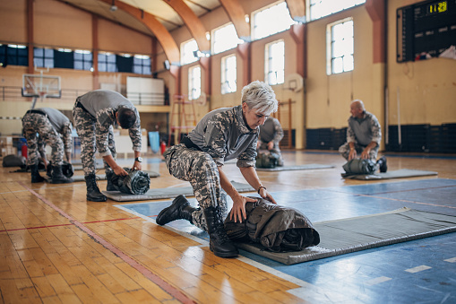 Diverse group of people, soldiers on humanitarian aid preparing sleeping bags for civilians in school gymnasium, after natural disaster happened in city.