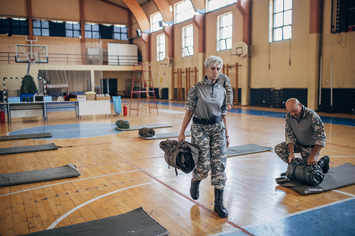 Diverse group of people, soldiers on humanitarian aid preparing sleeping bags for civilians in school gymnasium, after natural disaster happened in city.