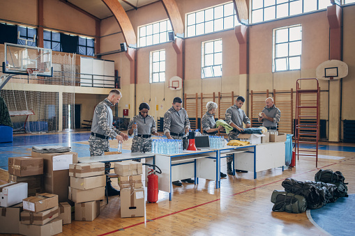Diverse group of people, soldiers on humanitarian aid preparing donations for civilians in school gymnasium, after natural disaster happened in city.