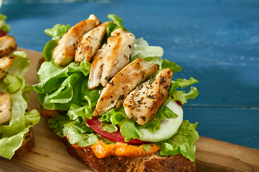 Tasty chicken and vegetable sandwiches with sauce on a wooden cutting board, on blue wooden home or restaurant table, close up photo with a space for copy