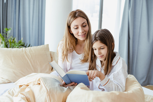 Mother and daughter sitting on the bed, mother is holding a book and reading it to her