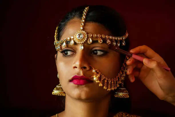 indian beautiful female in golden rich jewelery and tradition saree face closeup professional make-up wearing bindi on head .Artist doing to muslimgirl with bindis maang tikka ,nath,nose Pin