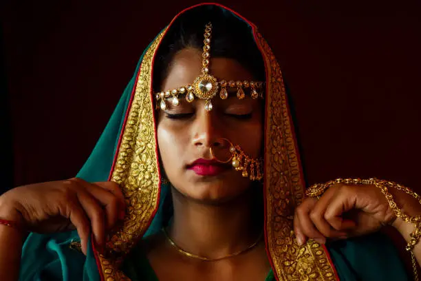 Photo of portrait indian beautiful female in golden rich jewelery and tradition saree face closeup professional make-up wearing bindi on head .Muslim Woman face portrait with bindis maang tikka ,nath,nose Pin