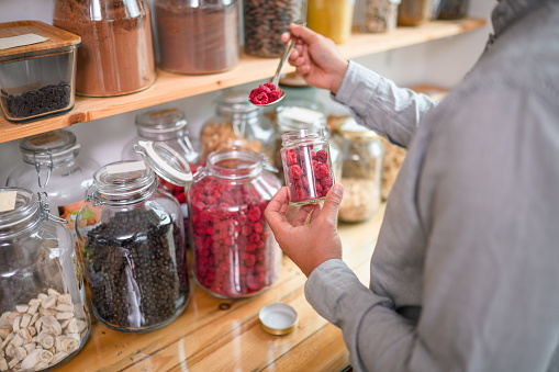 Asian mid adult male packing dried raspberries to a small mason jar in a sustainable grocery store.