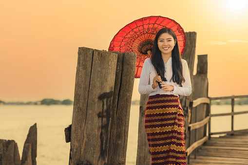 portrait of smiling Myanmar woman with red umbrella in Burmese traditional dress at U bein bridge Amarapura in Mandalay