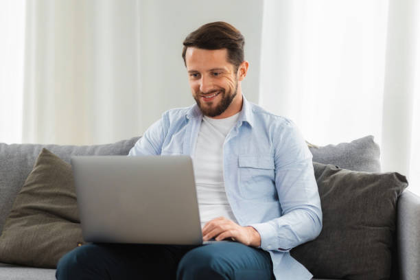 istock Man working or studying online remotely using laptop. Positive attractive caucasian male sitting on sofa at home using computer, smiling 1417831200