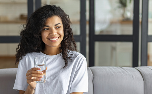 Healthy lifestyle concept. Beautiful young Afro American woman with glass of clean water, looking away and smiles friendly, copy space