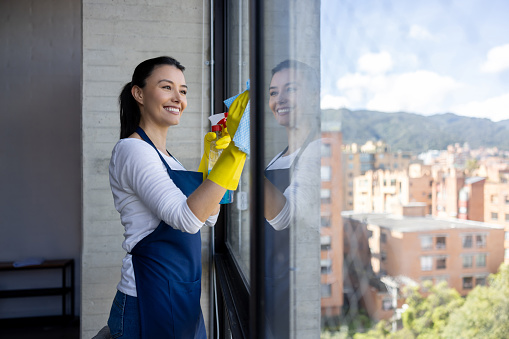 Happy professional cleaner cleaning a window at an apartment using a cleaning product for glass â housework concepts