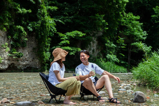 coppia di adulti che si rilassano sulle sedie da campeggio sorseggiando un drink in riva al fiume per un weekend di divertimento - solo giapponesi foto e immagini stock