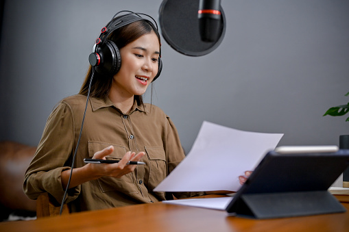 Talented young Asian female freelance blogger or radio host reading her podcast script and running her online radio channel in her studio.