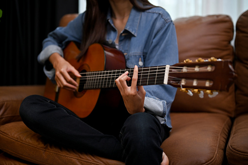 Attractive young Asian female playing her classic guitar in the living room, on her vintage brown leather sofa. cropped image