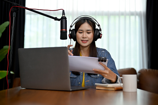 Charming Asian woman recording a podcast on her laptop with headphones and a microphone. Female podcaster making audio podcast from her home studio.