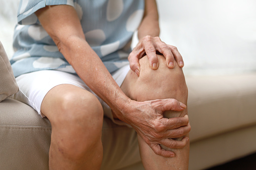 Una mujer asiática mayor masajeando su rodilla, sufriendo de dolor en la pierna mientras estaba sentada en el sofá de la sala de estar de su casa. photo