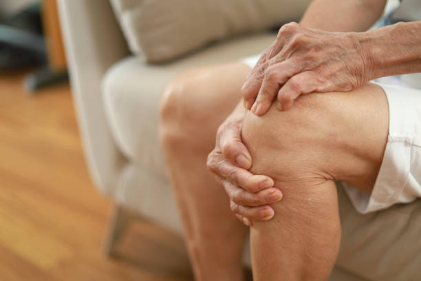 自宅のリビングルームのソファに座りながら、膝をマッサージするアジアのシニア女性 - arthritis senior adult rheumatoid arthritis sadness ストックフォトと画像