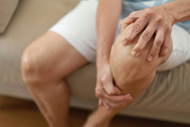 starsza azjatka masująca kolano, cierpiąca na ból nogi podczas siedzenia na kanapie w salonie w domu. widok pod dużym kątem - arthritis senior adult rheumatoid arthritis sadness zdjęcia i obrazy z banku zdjęć