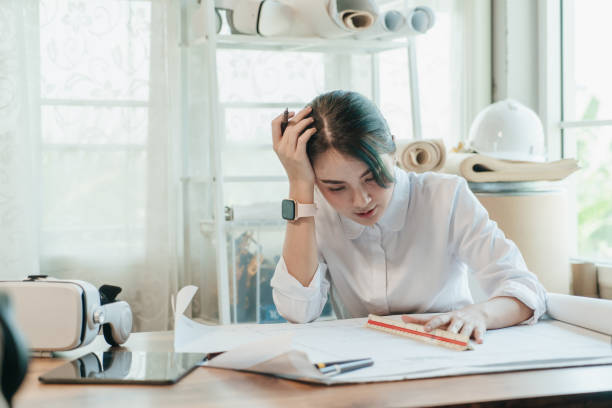 istock Stressed Asian woman architect having headache while working on blueprint in office. 1417845993