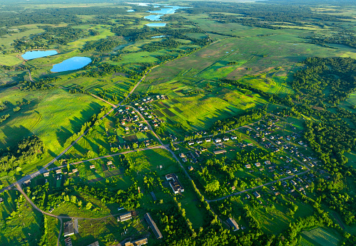 Village at lake, aerial view. Country houses at river in countryside. Roofs of country wooden houses near a pond in village. Green farm fields and forests in rural landscape. Farmland, Agriculture.