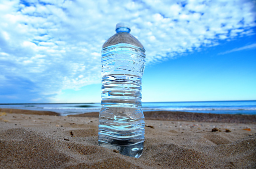 Plastic bottle with mineral water on beach by sea. PET bottle on the sand. Plastic trash thrown into the ocean. Thirst for water during dry and hot weather. Clean water in bottle at coastline.