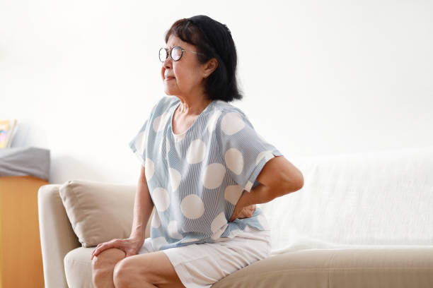 пожилая азиатка, страдающая от болей в спине, сидит дома на диване - backache pain women illness стоковые фото и изображения