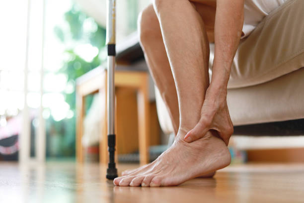 Senior woman sitting on sofa holds her ankle injury, feeling pain. stock photo