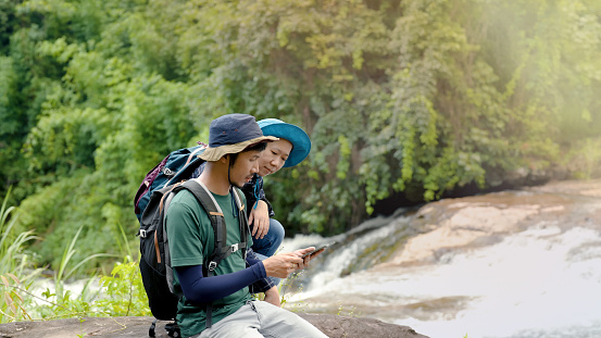 Asian man is suggesting his female partner map from a tablet to the next hiking and trekking route after admiring the beautiful waterfall on their vacation. Tourist couples love climbing and trekking.