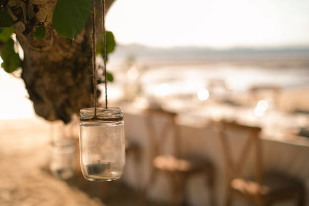 Close up the jar candle hanging on the tree with long table wedding dinner setup on the beach at Thailand in the evening. Wedding party concept. Decoration outdoor restaurant at the beach.
