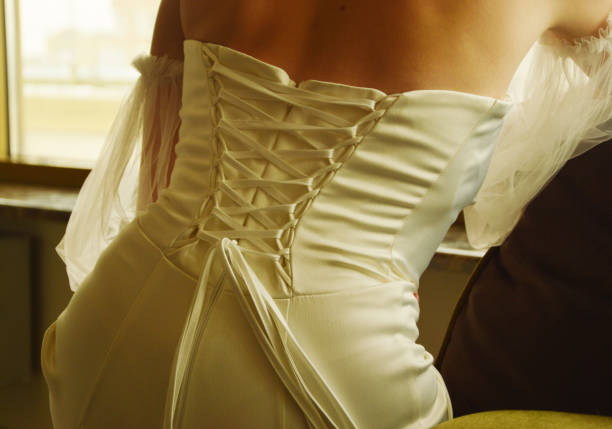 The bride in a beautiful wedding dress sits on a chair. View from the back. Corset lacing is an element of a wedding dress.