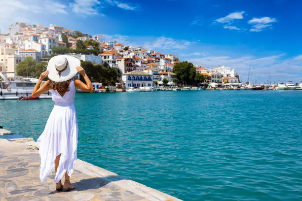 A beautiful tourist woman in a white summer dress enjoys the view to the city of Skopelos island, Sporades, Greece