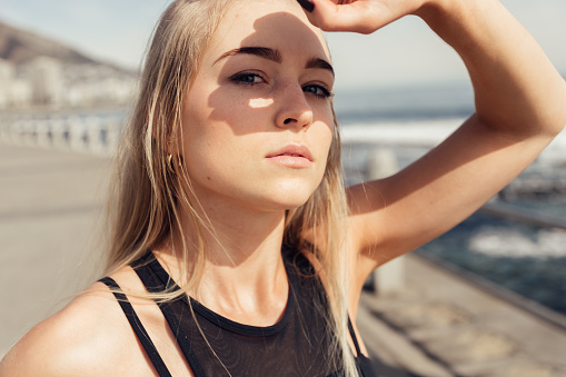 Close-up portrait of confident young woman shielding eyes with hand while exercising outdoors on sunny day