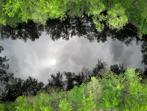An aerial view of a river with reflections of grey clouds and surrounded by trees
