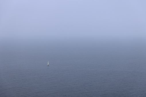 White sailing yacht glides on metal color water of the Tyrrhenian sea, which merges on the horizon with the sky of the same color, Province of Livorno, Island of Elba, Italy