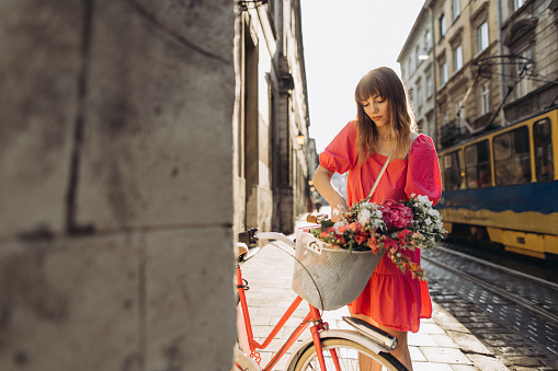 A beautiful woman with the bicycle with flowers in the basket in the city street in the morning