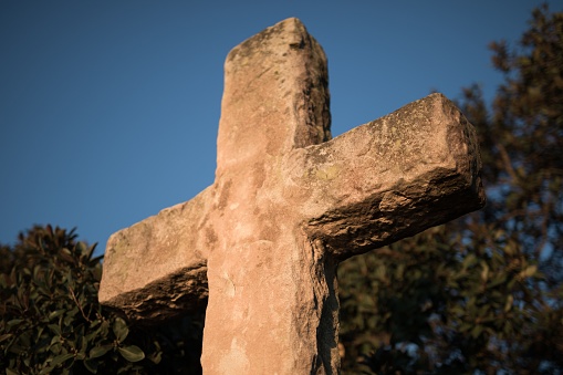 A low angle of a stone cross on a pedestal against green trees at golden hour